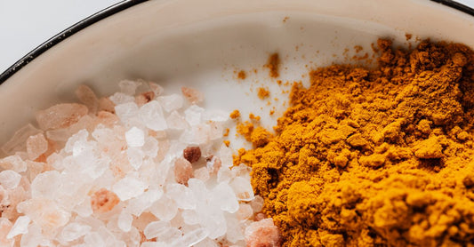 A small glass jar of fine white vitamin C crystals on a timber surface with a stainless steel scoop, illustrating the vitamin C crystals vs tablets debate.